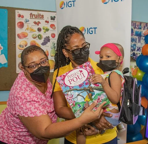 (L-R) Leslie Ann Baxam, Draw Coordinator- Lead at IGT and Chevaughn Joseph, Co-Founder of JBF, joyfully handed a signed copy of the local children’s book, Petra and the Poui by Phillip Simon to Adira, to a patient at the pediatric oncology clinic.