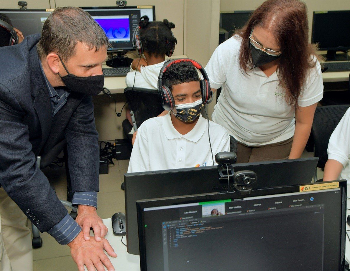 (L-R) Mr. Brendan Hames, IGT Caribbean Regional Director, Ms. Sita Persad - Executive Director at the Cotton Tree Foundation assists one of the students during a session of the second staging of IGT’s Coding and Robotics Rock! Camp.